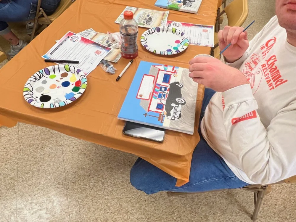 A person sitting at a table painting a scene of a storefront with a truck in front. The table is covered with a brown tablecloth and has paint palettes, brushes, flyers, and a water bottle on it.