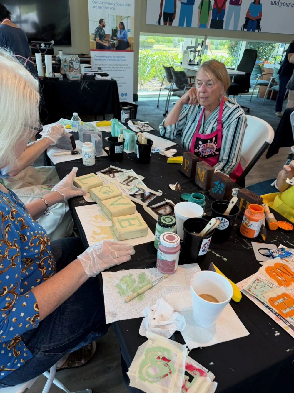 Two women sit at a table engaged in craft activity, painting and decorating wooden blocks. The table is scattered with paint supplies, brushes, and paper towels. One woman wears a striped shirt and pink apron, the other wears a blue patterned top, bo