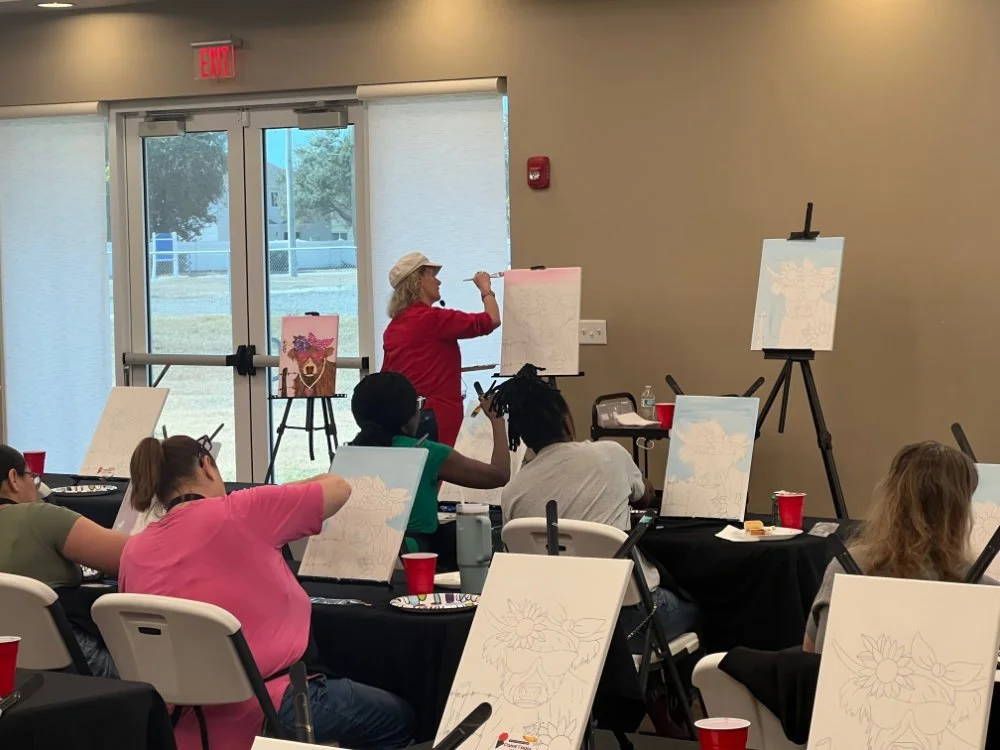 A woman in a red shirt and hat is leading a painting class in a room with large windows. Students are seated at tables with canvases, some of which are in progress, and are listening to her demonstration.