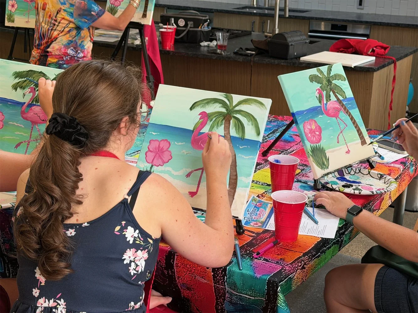 People painting tropical beach scenes with pink flamingos, palm trees, and hibiscus flowers on canvas during a painting class.