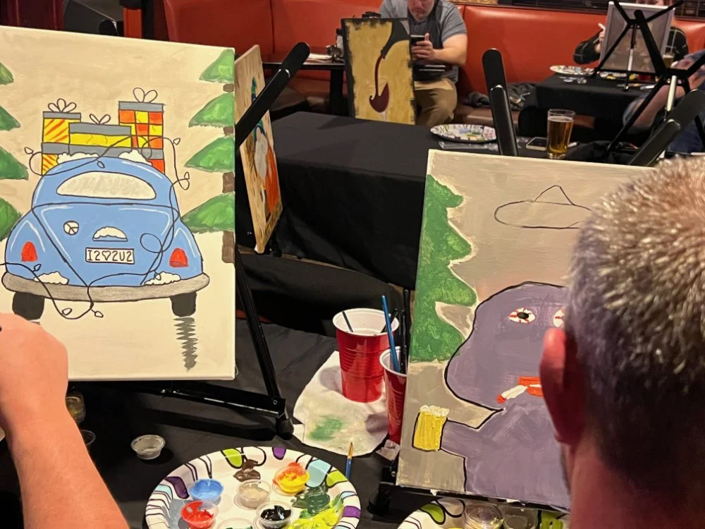 People painting colorful canvases at an art event, with some completed paintings of a blue car with gifts and a purple creature holding a yellow cup, surrounded by painting supplies on the table.