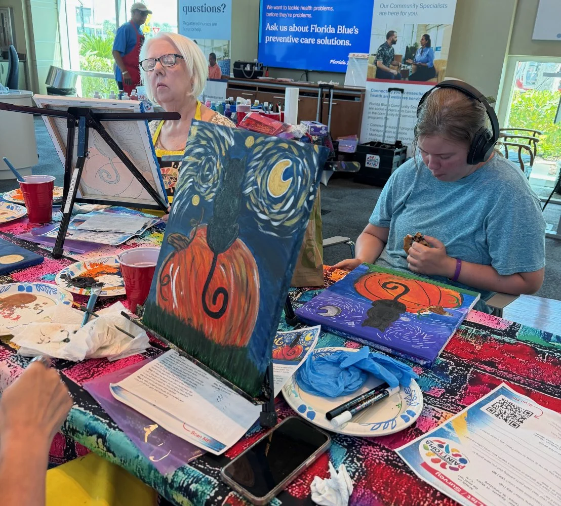 People painting Halloween-themed pictures, one with a black cat and pumpkin, at an indoor event with art supplies and informational posters.