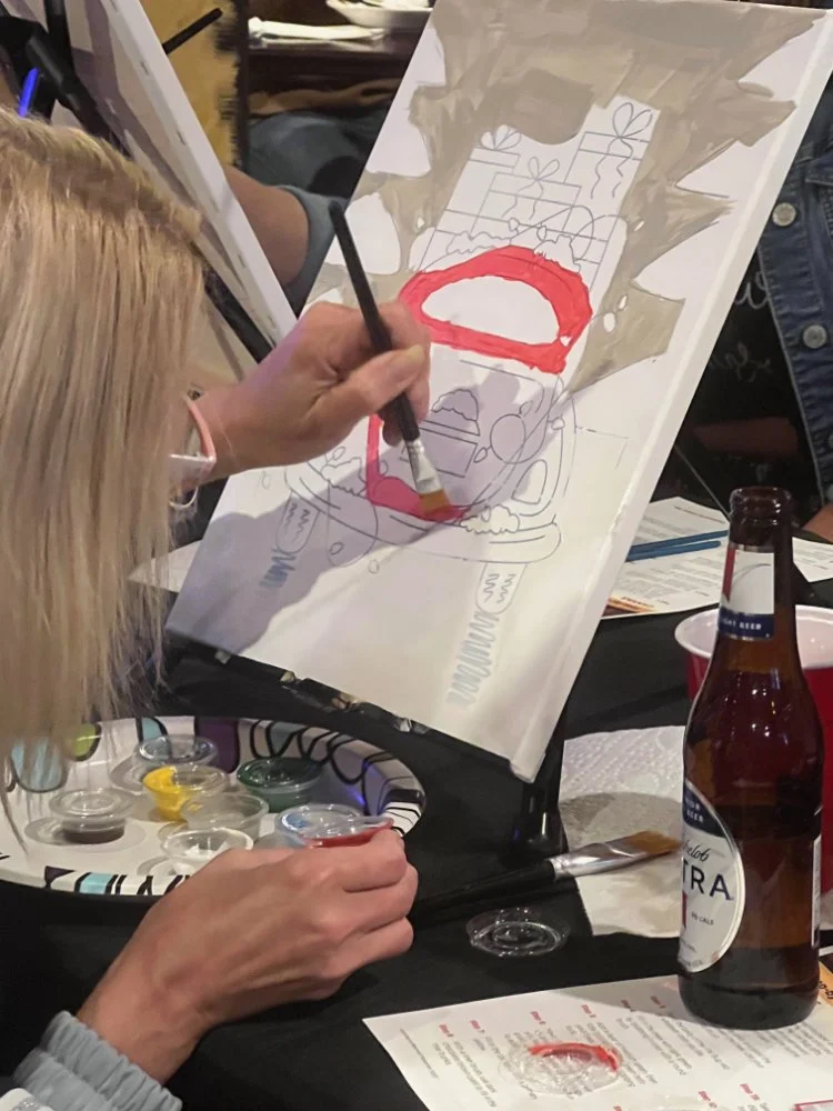 Person using paint and a brush to create a holiday-themed painting, featuring a red Santa face and Christmas presents, at a table with paints, a beer, and a paper napkin.