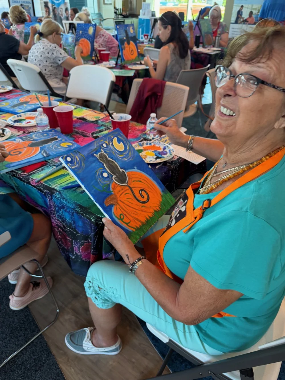 A woman smiling while holding a colorful painting of a black cat sitting on an orange pumpkin, participating in a group painting class with other people at a table covered with art supplies.