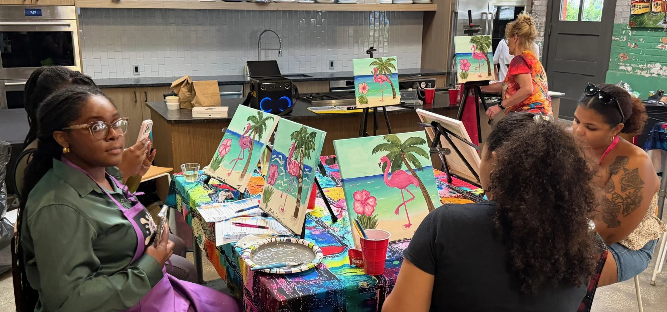 Group of women participating in a painting class, with tropical-themed canvases featuring flamingos, palm trees, and flowers, in a colorful studio with art supplies on a table.