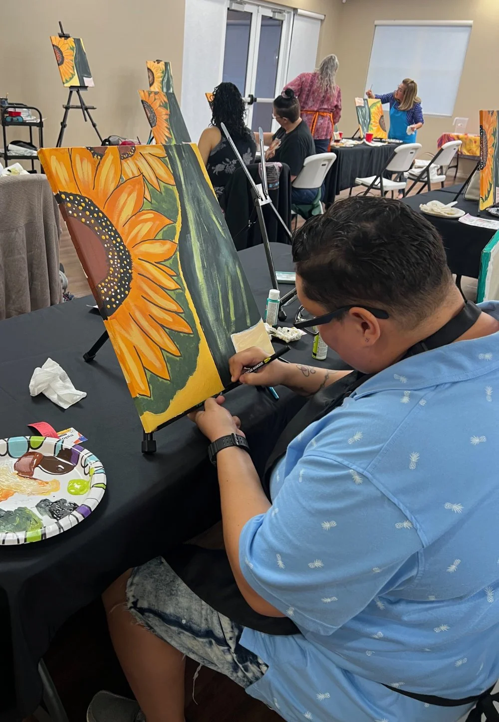 A man painting a sunflower on a canvas during a group painting class in a room with other participants, some of whom are working on their own paintings.
