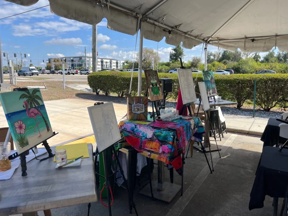 Outdoor art booth under a tent displaying colorful paintings on easels, including a flamingo, a character, and a house with a palm tree in a parking lot.