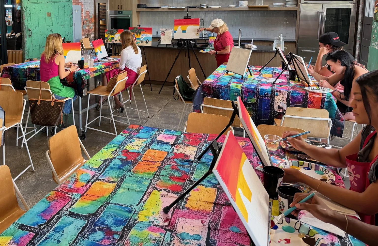 People participating in an outdoor painting class with colorful abstract paintings, tables covered with vibrant art-filled tablecloths, and a woman instructing at an easel.
