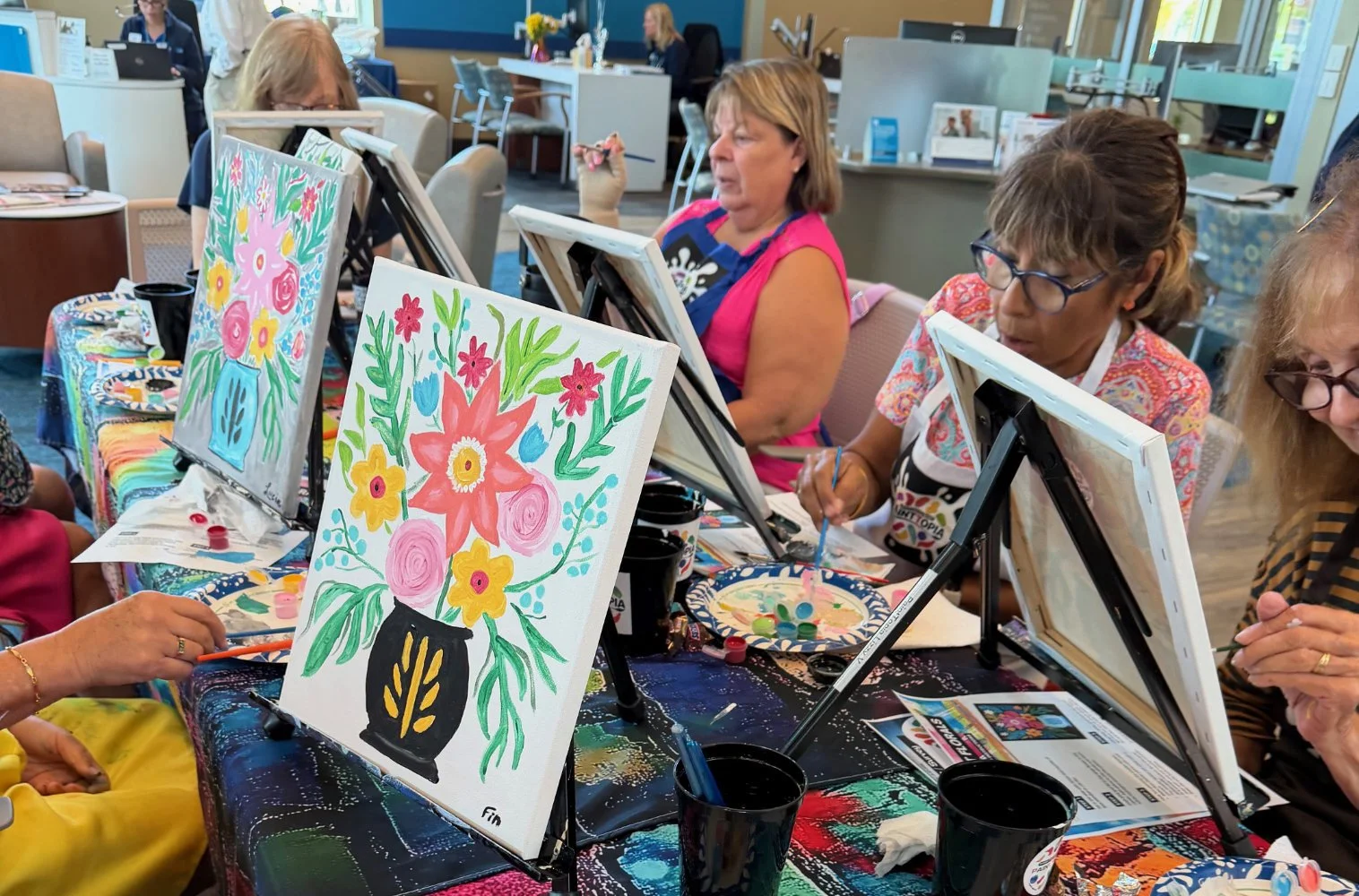 Women and children participating in a painting class at a colorful indoor art studio, painting floral designs on canvases.