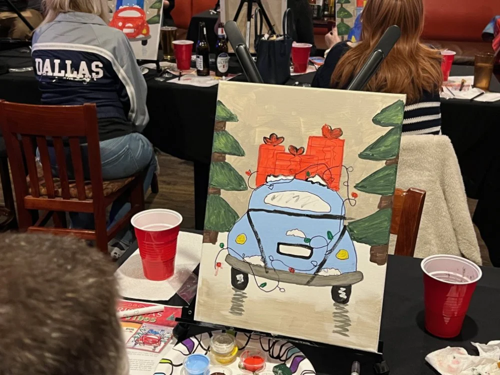 A painting of a blue car with Christmas lights wrapped around it, surrounded by green trees and red Christmas presents in the background, on a table in a busy indoor setting with people and drinks.