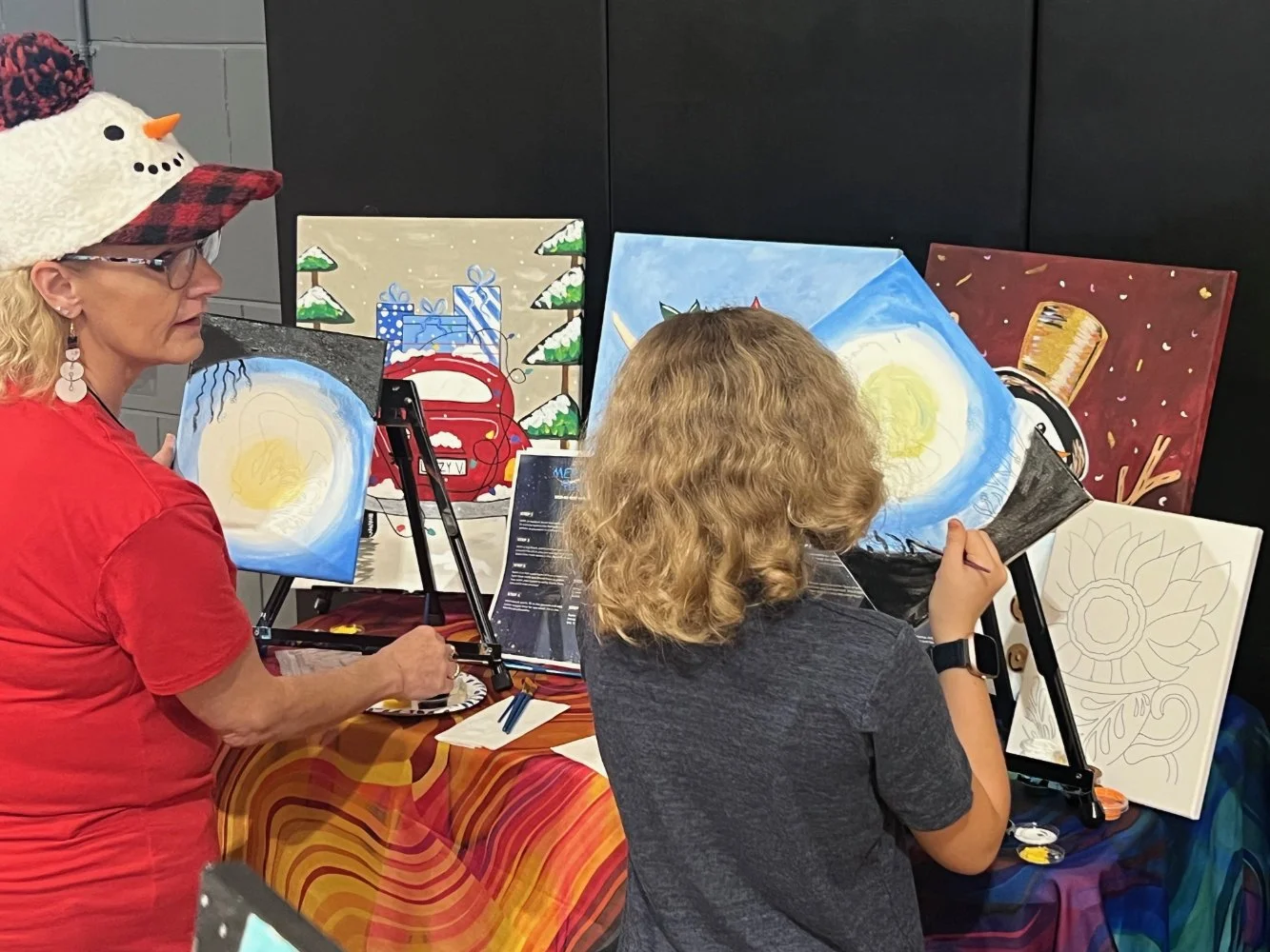 A woman in a red shirt and snowman hat and a girl with curly blonde hair are painting on canvases at an art event, with multiple holiday-themed paintings displayed behind them.