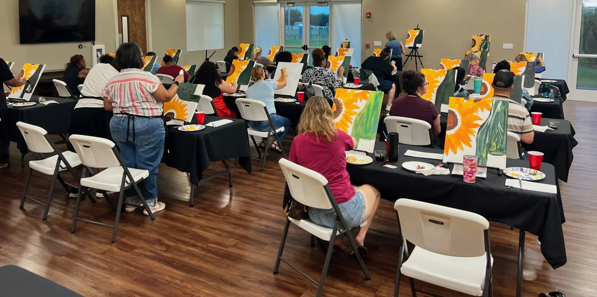 A group of people participating in a painting class in a room with large windows, large sunflower paintings on easels in front of them, and a painting instructor at the front.