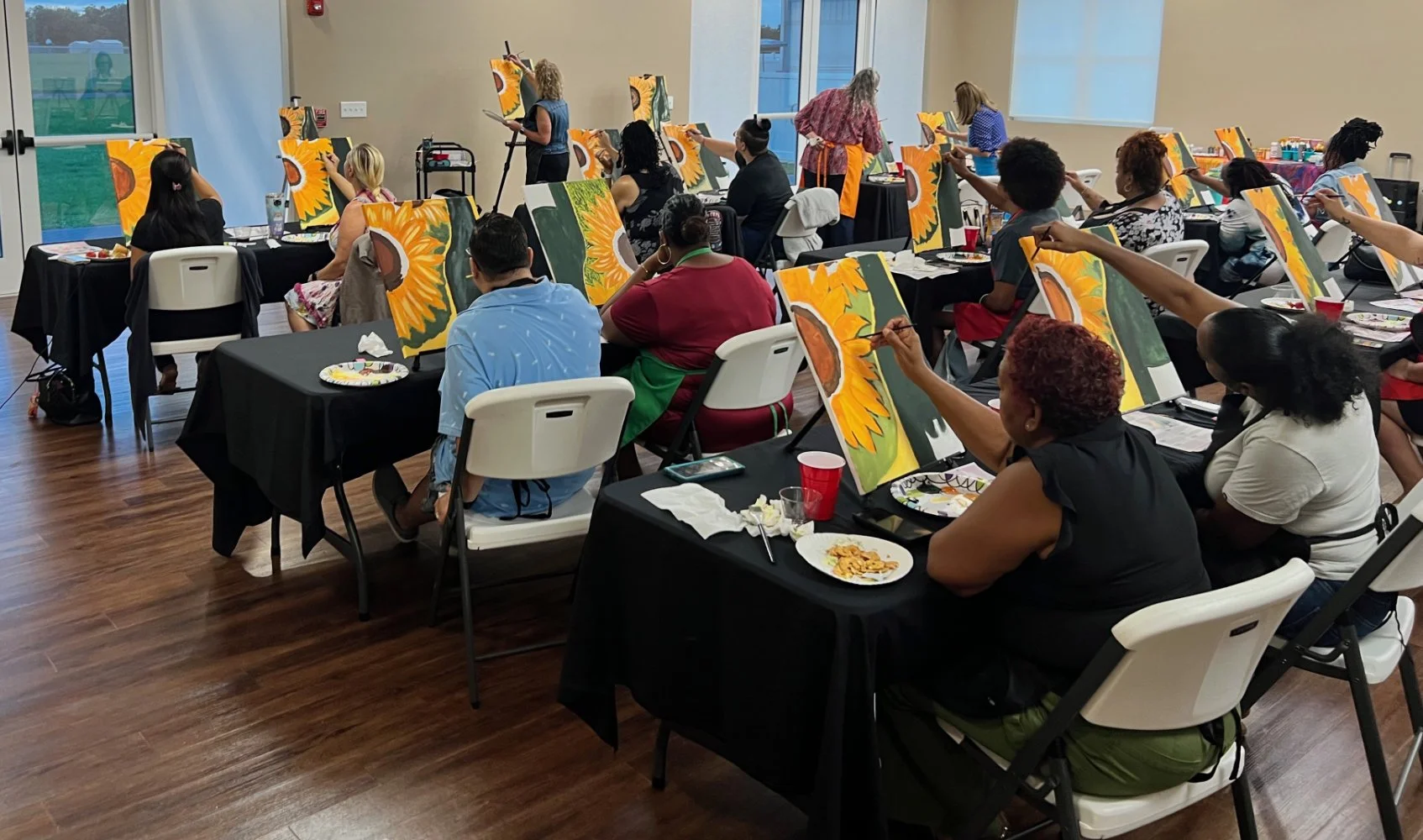 People participating in a group painting class, each working on a sunflower artwork at tables in a well-lit room.