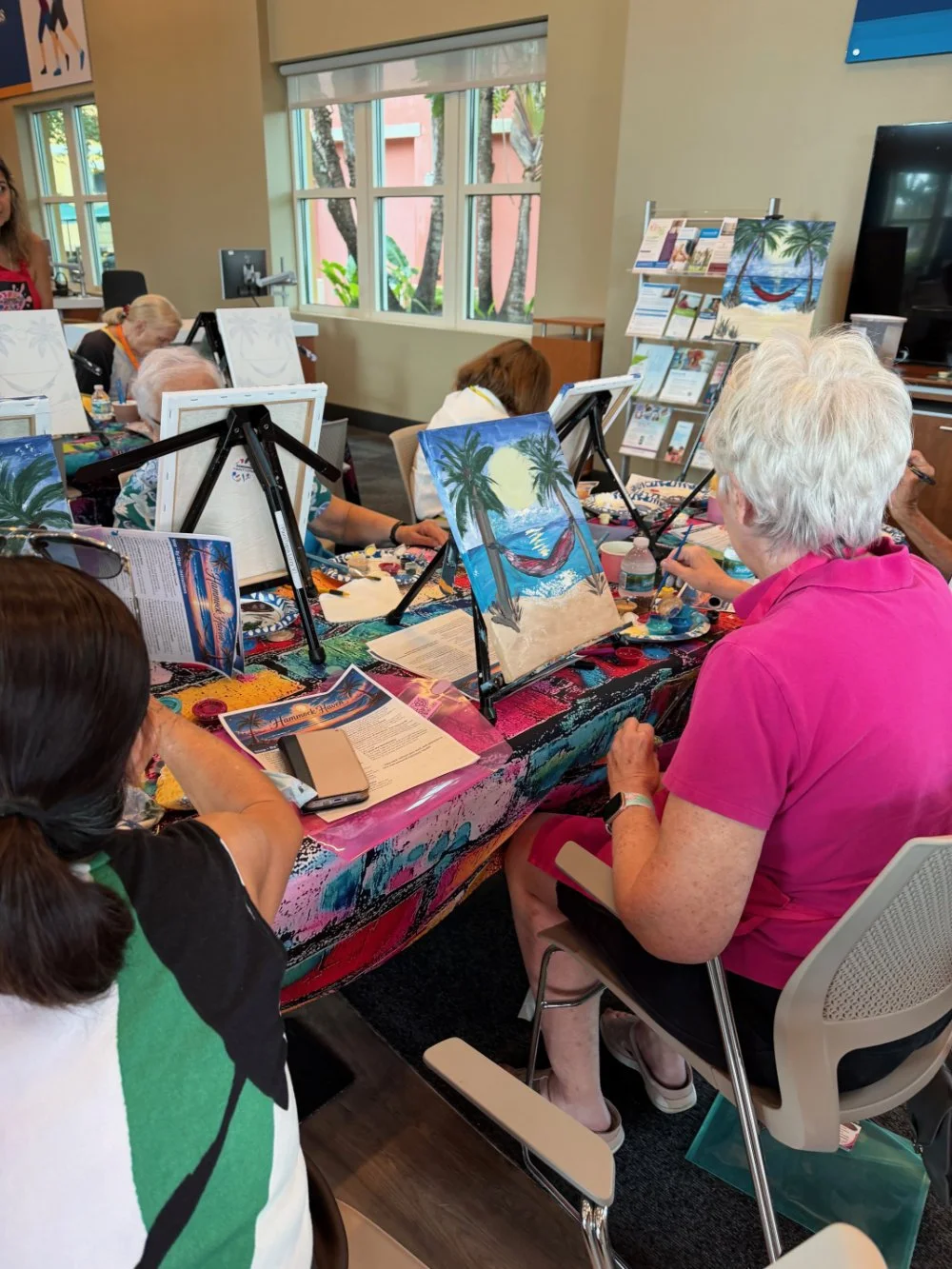 People painting tropical beach scenes with palm trees on canvas at a group art class.