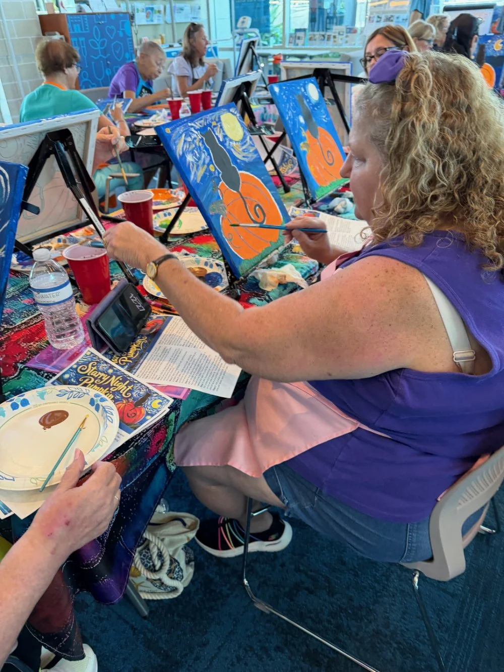 Group of women participating in a painting class, creating pictures of a black cat with a full moon and orange swirl background, seated at tables with painting supplies.