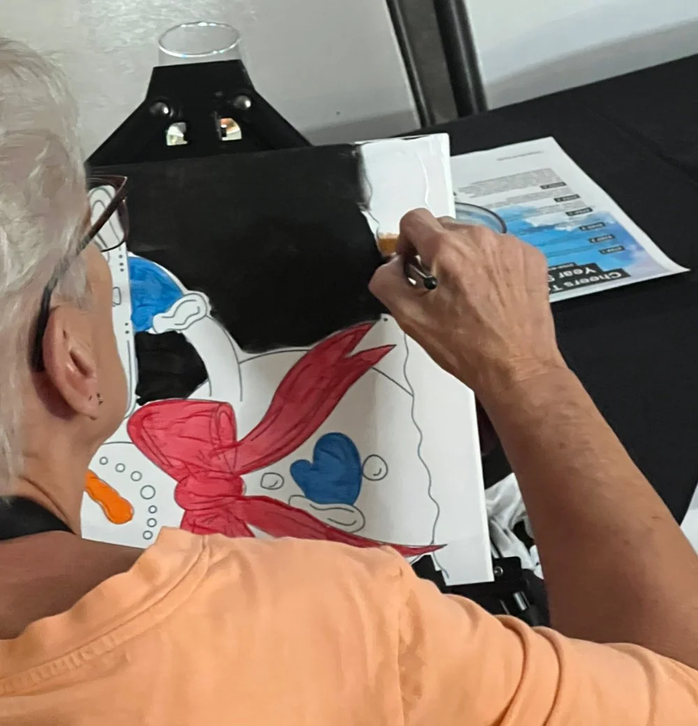An elderly woman with glasses coloring a large drawing of a cartoon mermaid on paper. She is using markers or crayons to fill in the colors, with a black and white background and a blue heart on the drawing. The table has some printed materials and a