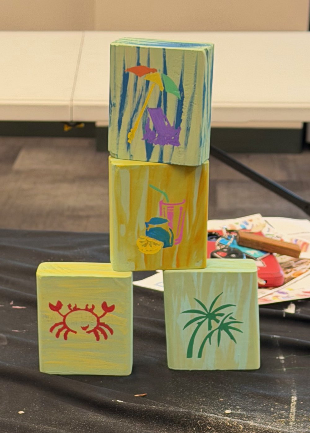 Four painted wooden blocks arranged in a pyramid with summer-themed drawings, including an umbrella, a drink with a straw, a crab, and palm trees, on a black surface with art supplies in the background.