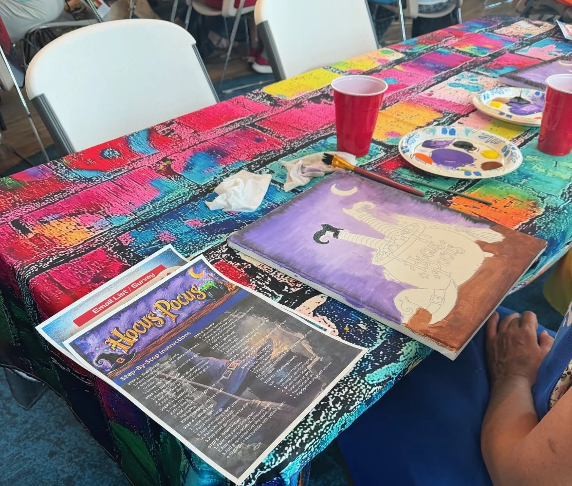 A table covered with a colorful, painted tablecloth hosting painting supplies, disposable cups, and a painting of a swords and the moon with the words 'Horus Rocus.' There are chairs and other tables in the background.