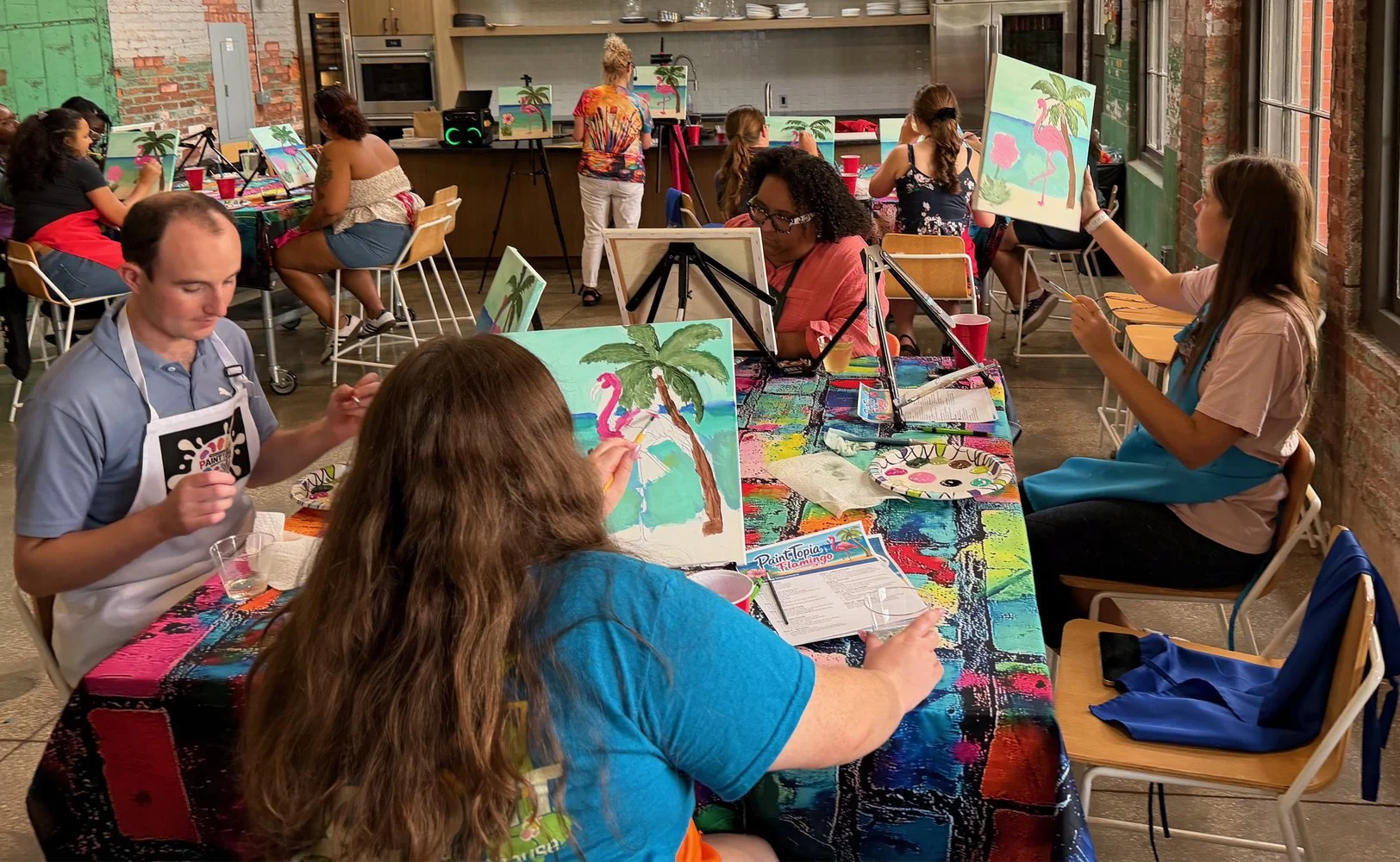People participating in a painting class, creating tropical-themed artwork with pink flamingos, palm trees, and ocean scenes. The room has exposed brick walls and large windows, and the tables are covered with colorful, paint-stained tablecloths.
