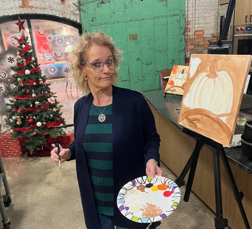 A woman with glasses, curly blonde hair, wearing a navy blazer over a striped teal and black shirt, stands next to an easel with a painting of a white pumpkin. She holds a paintbrush in one hand and a paper plate with paint in the other. Behind her i