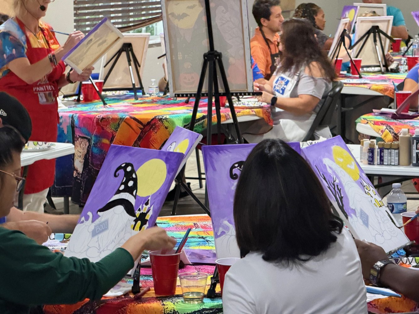 People participating in a painting class, working on Halloween-themed paintings of a black cat against a yellow moon, with some paintings in progress and art supplies on the tables.