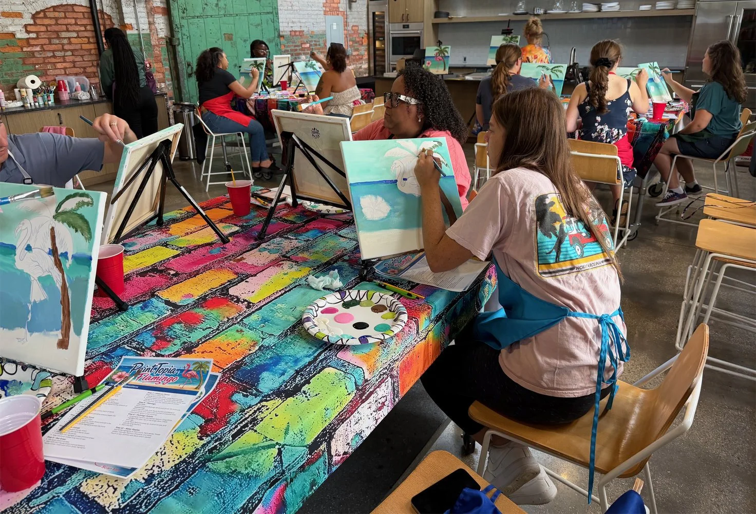 Group of people participating in a paint and sip class, painting tropical-themed canvases featuring palm trees and swans in a brightly lit, industrial-style studio with brick and green walls, with art supplies and drinks on the colorful table.