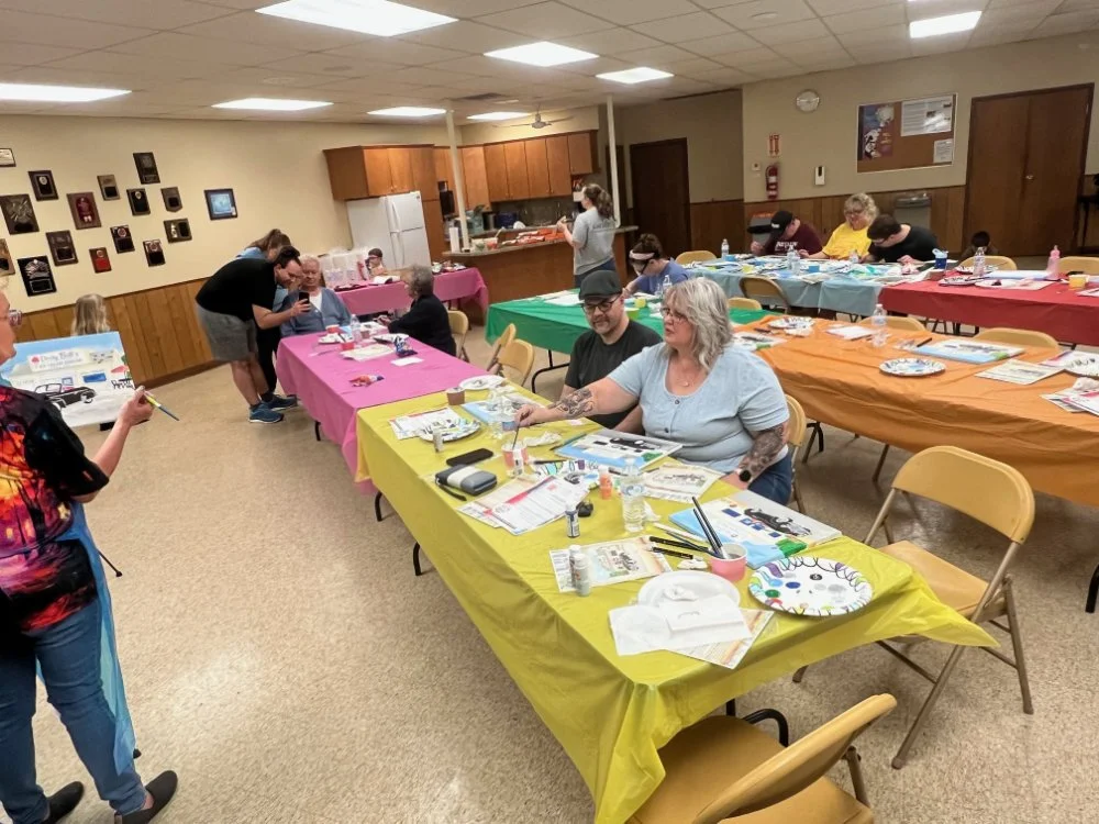 Community center room with decorated tables covered in colorful tablecloths, where people are painting and crafting for a group activity or class. Participants are engaged in creative activities with art supplies, and the background shows a kitchen a