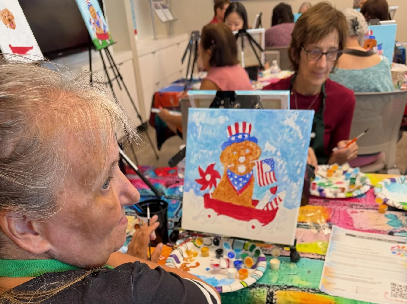 People painting patriotic-themed pictures of a gingerbread man with an American flag, on small canvases, at an art event. The focus is on a person with gray hair and a black shirt in the foreground, with others working on their art in the background.