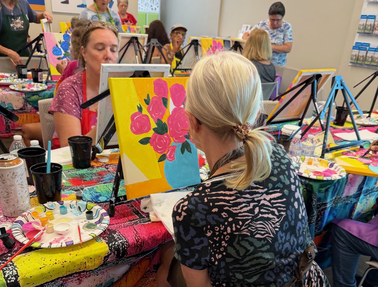 People participating in a paint and sip class, painting pink flowers in a blue vase on bright yellow and orange backgrounds at a table set with painting supplies, while others work on similar paintings around the room.