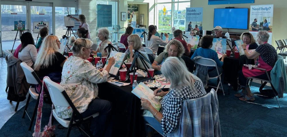 A group of people participating in a painting class inside a well-lit room with large windows, some are painting on canvases and others watching a teacher, with signs about health questions and help in the background.