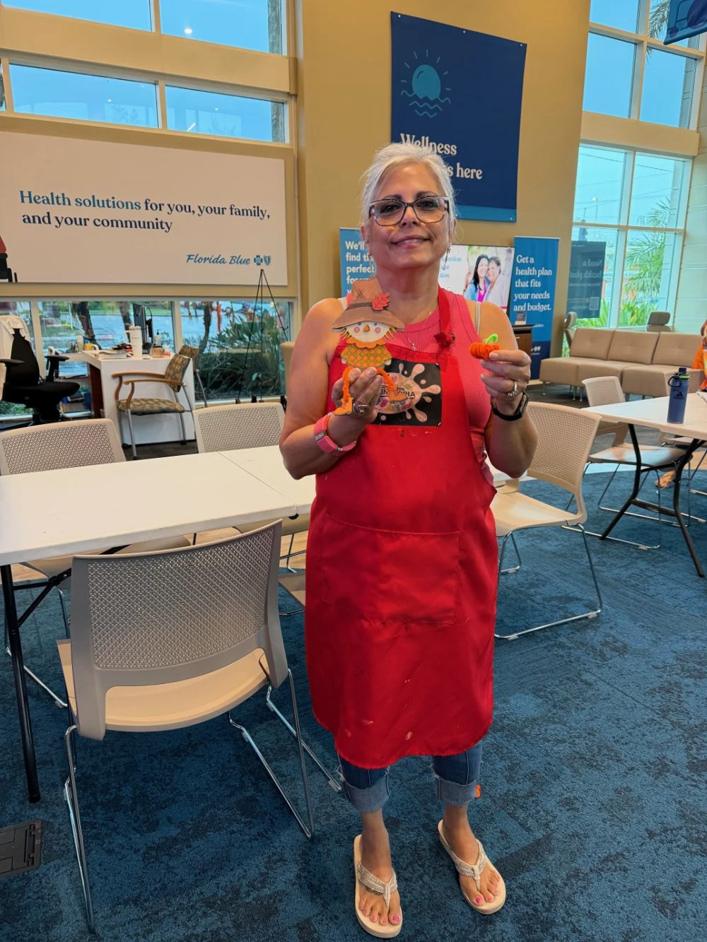 Woman in a red apron standing indoors, holding a small decorative turkey figure and a carrot, with large windows and posters in the background.