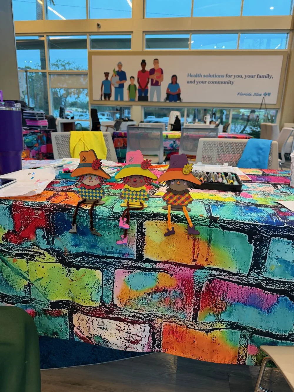 Colorful table with three paper craft dolls wearing witch hats, surrounded by art supplies in a community center or classroom with large windows and a Banner in the background.