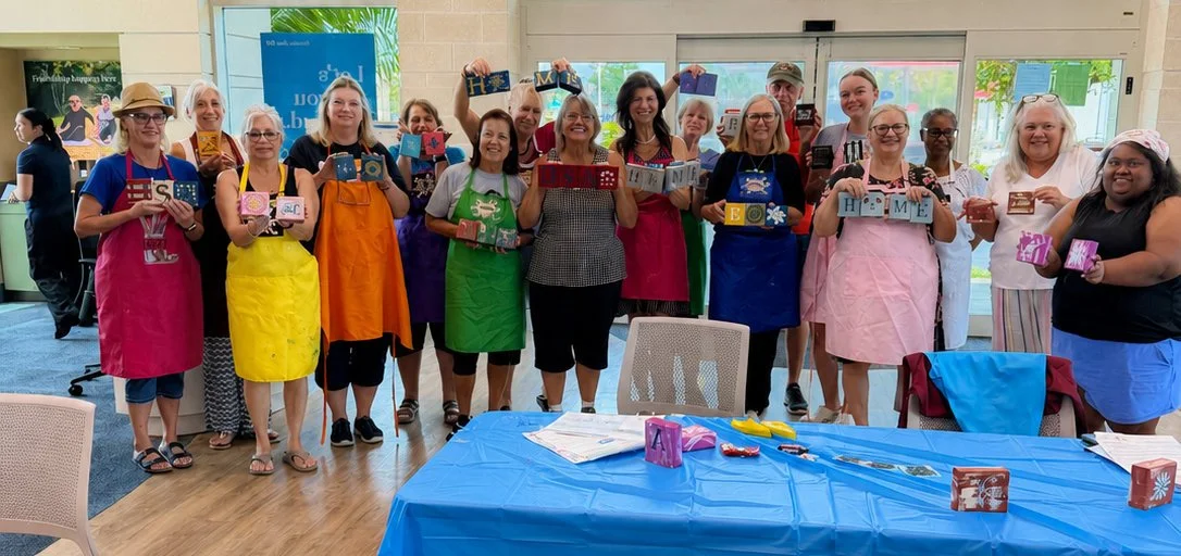 Group of women in colorful aprons holding decorated boxes in an indoor space with large windows.