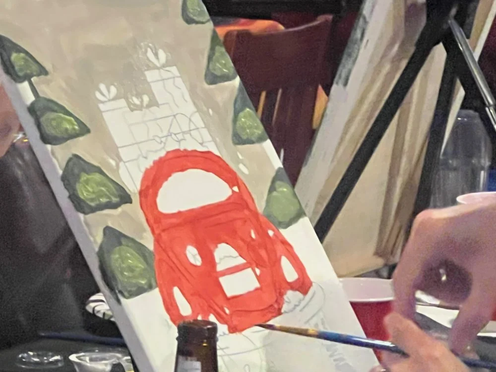 A painting in progress of a red backpack surrounded by green leaves, with a hand holding a paintbrush working on the artwork.
