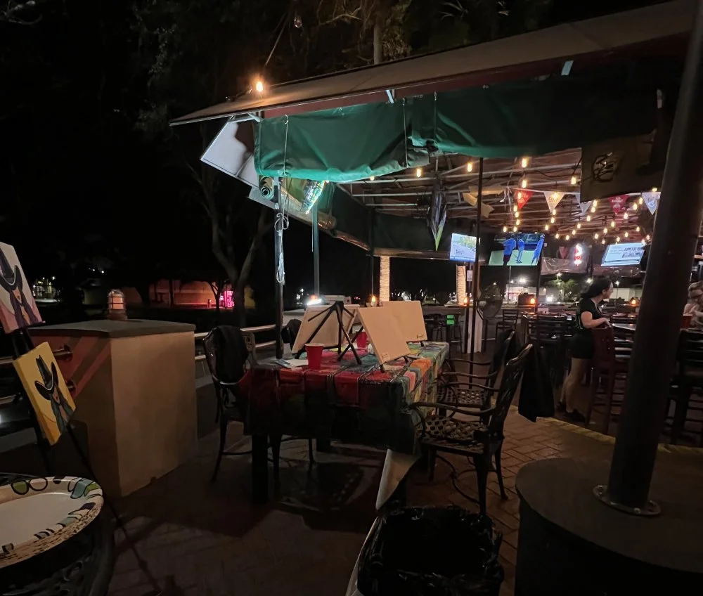 Nighttime outdoor art display setup at a bar or restaurant with paintings, easels, string lights, and a TV screen in the background.