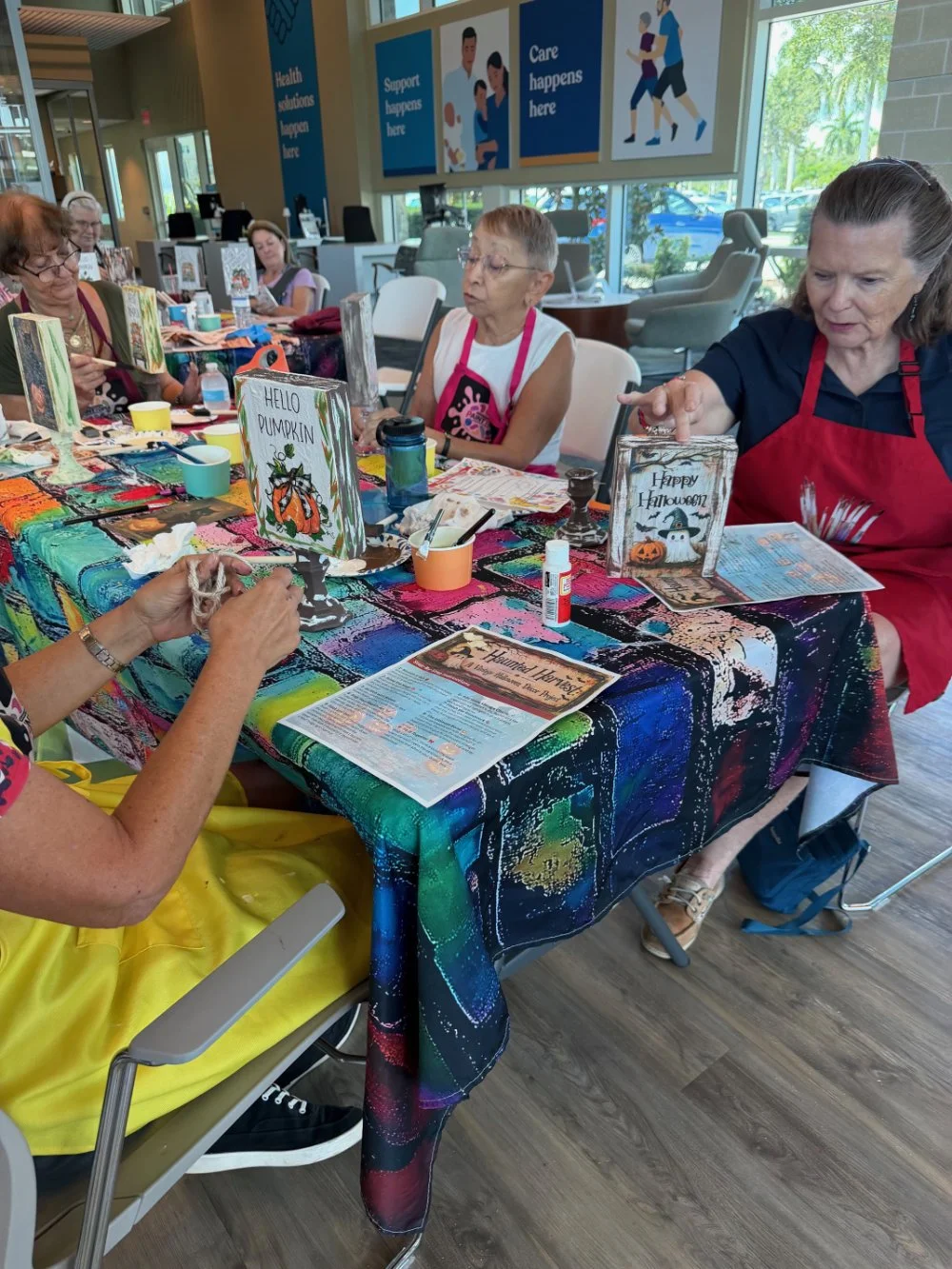 Group of elderly women participating in a Halloween-themed crafting activity at a community center, sitting around a colorful table with Halloween signs and decorations.
