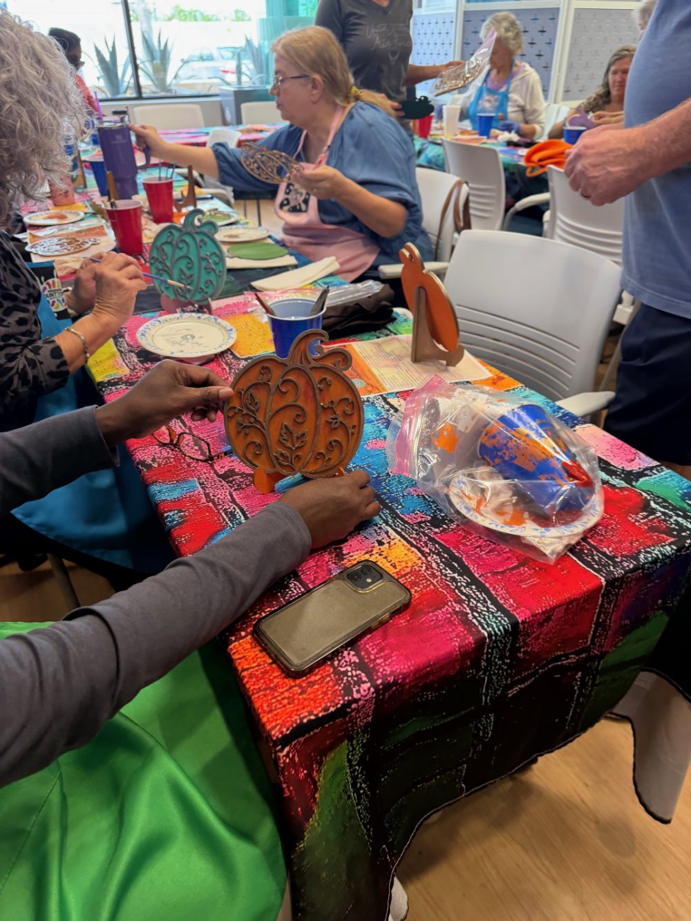 People gathered around a table decorated with a colorful tablecloth, painting and decorating carved wood pumpkin ornaments, with art supplies and disposable cups on the table.