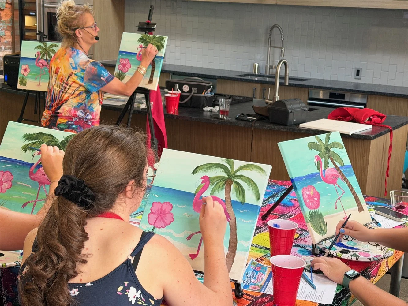 People participating in a paint and sip class, painting tropical beach scenes with pink flamingos, palm trees, ocean, and hibiscus flowers on canvases.