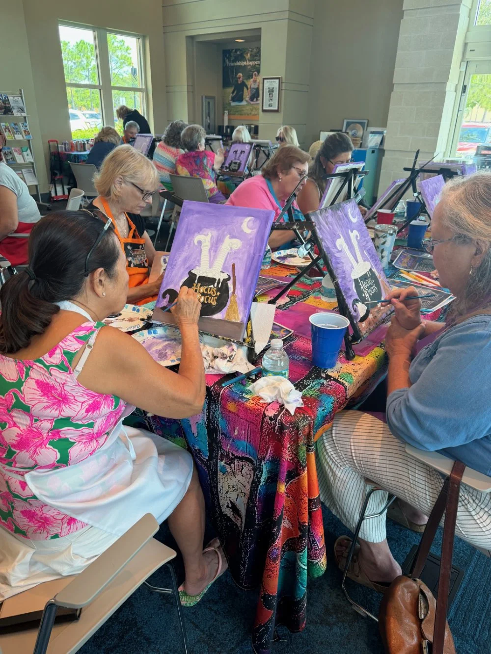 A group of women at a painting class, working on Halloween-themed paintings of a cauldron with a purple, night sky background on easels.