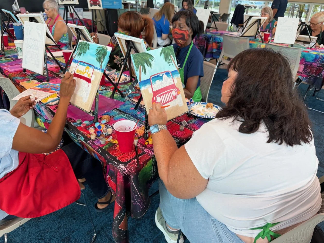 People participating in a painting class, painting beach and palm tree scenes on canvas, seated around a table covered with a colorful, splattered cloth, with supplies like paint bottles, brushes, and drinks visible.