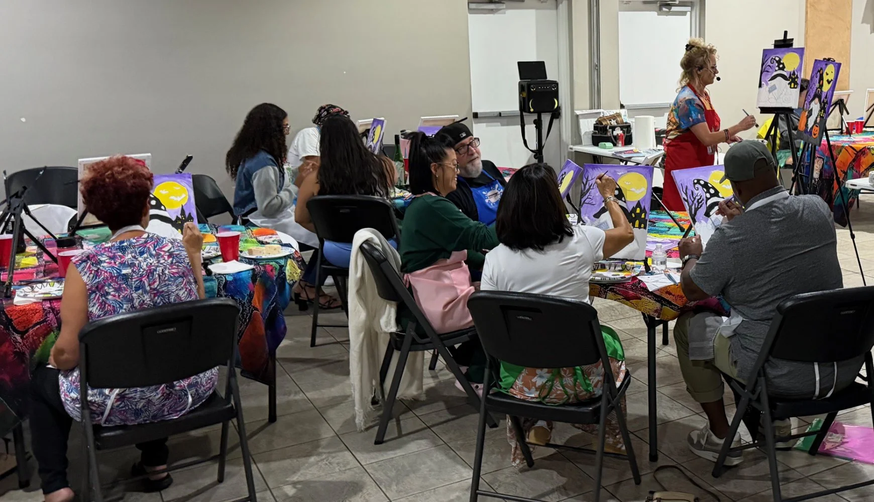 People participating in a Halloween-themed painting class, with some canvas paintings featuring spooky scenes, colorful tablecloths, and art supplies.