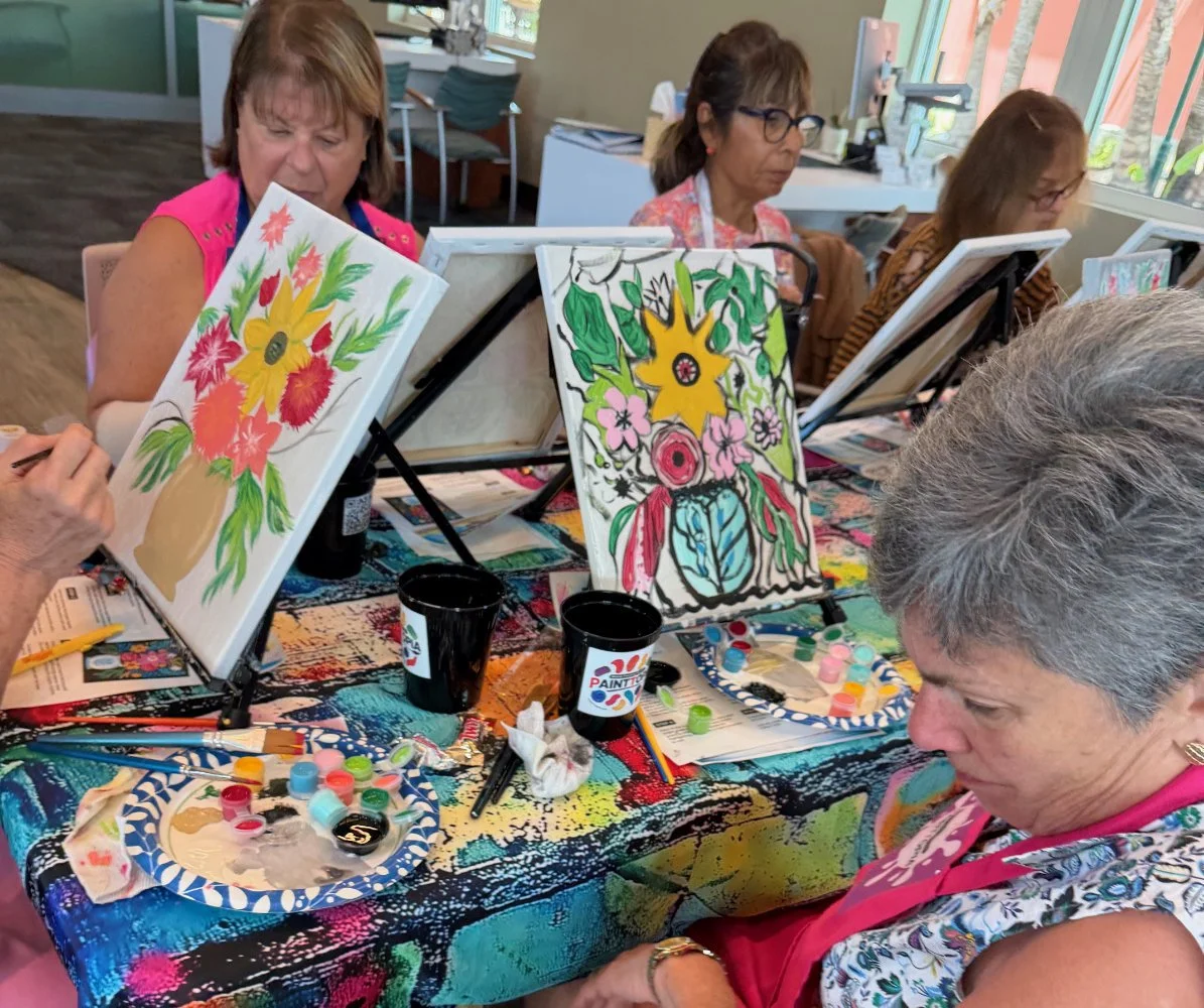 Group of women painting colorful floral artwork on canvases at a painting class, with paints and brushes on the table.