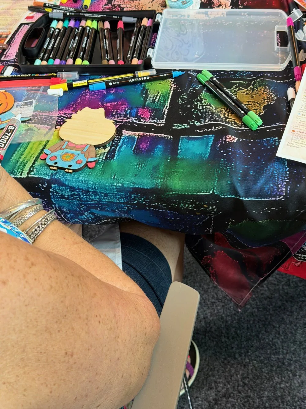 A table covered with markers, pens, and craft supplies, including a wooden craft figure, a carved wooden pendant, and a clear plastic container, with a colorful tablecloth underneath.