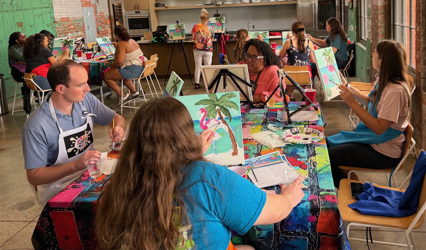 People participating in an indoor group painting class, creating tropical-themed artworks featuring palm trees and pink flamingos, with an instructor guiding them.