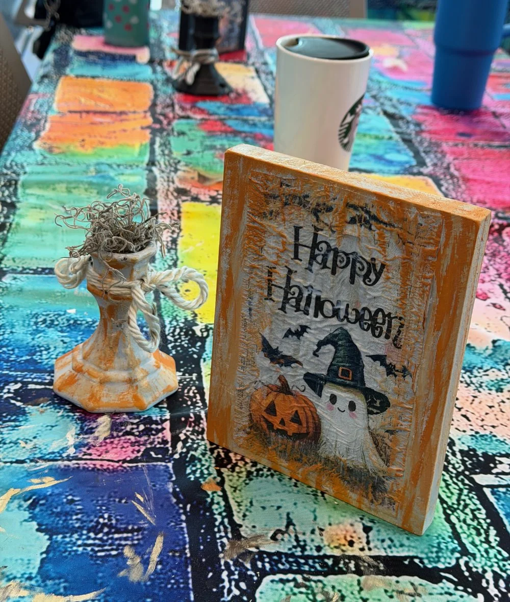 Decorative Halloween table setting with a colorful tablecloth, a white Starbucks cup, a small vase with dried flowers, and a Halloween sign featuring a smiling ghost, a witch hat, a pumpkin, and bats.