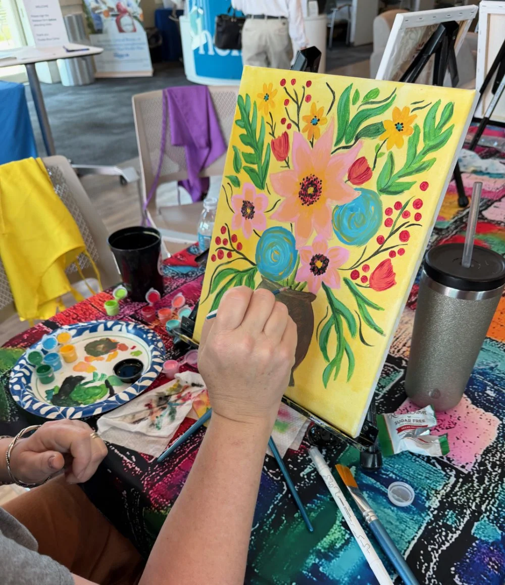 A person painting a colorful floral picture on a canvas at an art event, with various paints, brushes, and a water tumbler on the table.