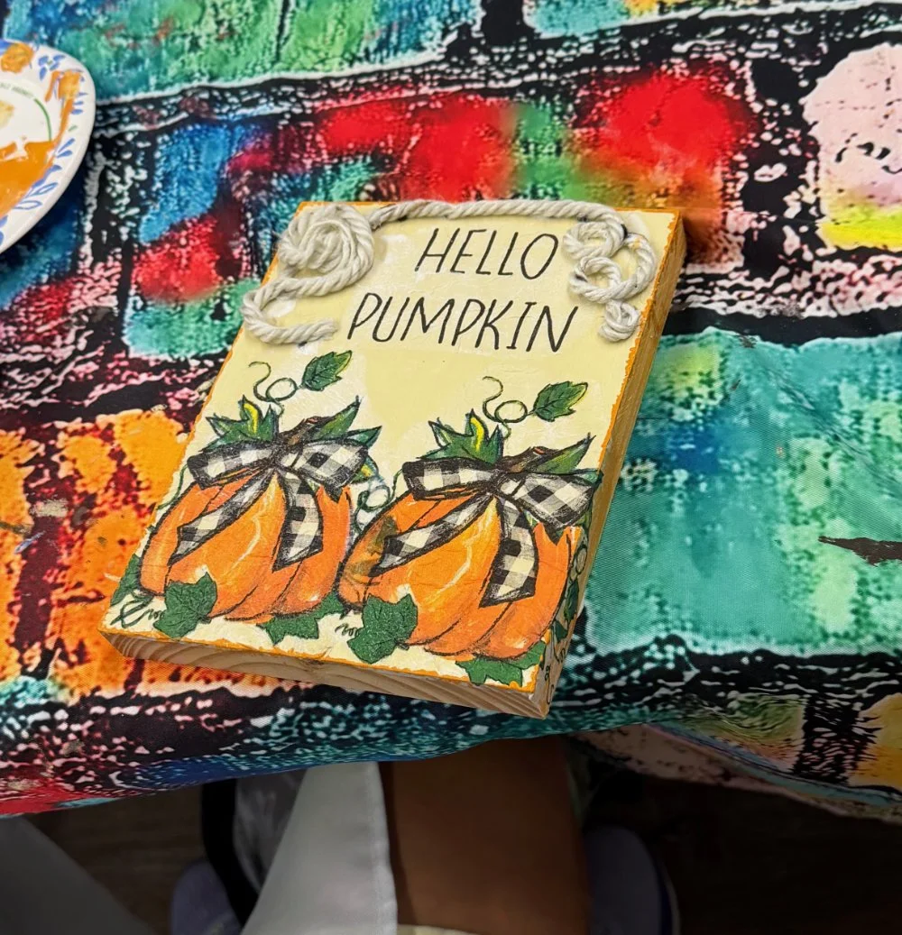 A decorated sign with the words 'Hello Pumpkin' above two illustrated pumpkins with black and white checkered bows, placed on a colorful, patterned tablecloth.