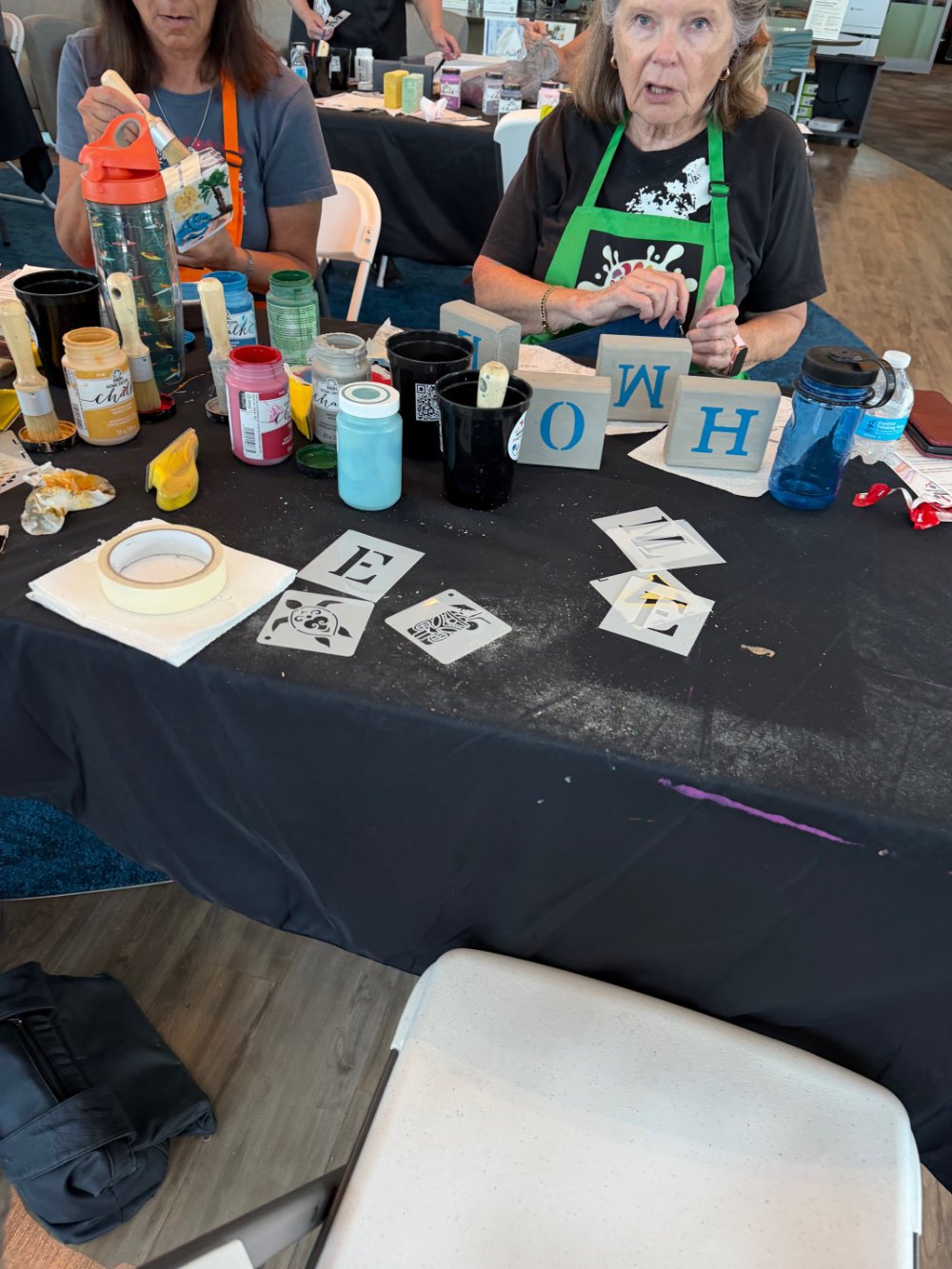 Women participating in an arts and crafts activity at a table with paints, brushes, signs spelling 'HOME', and various craft supplies.