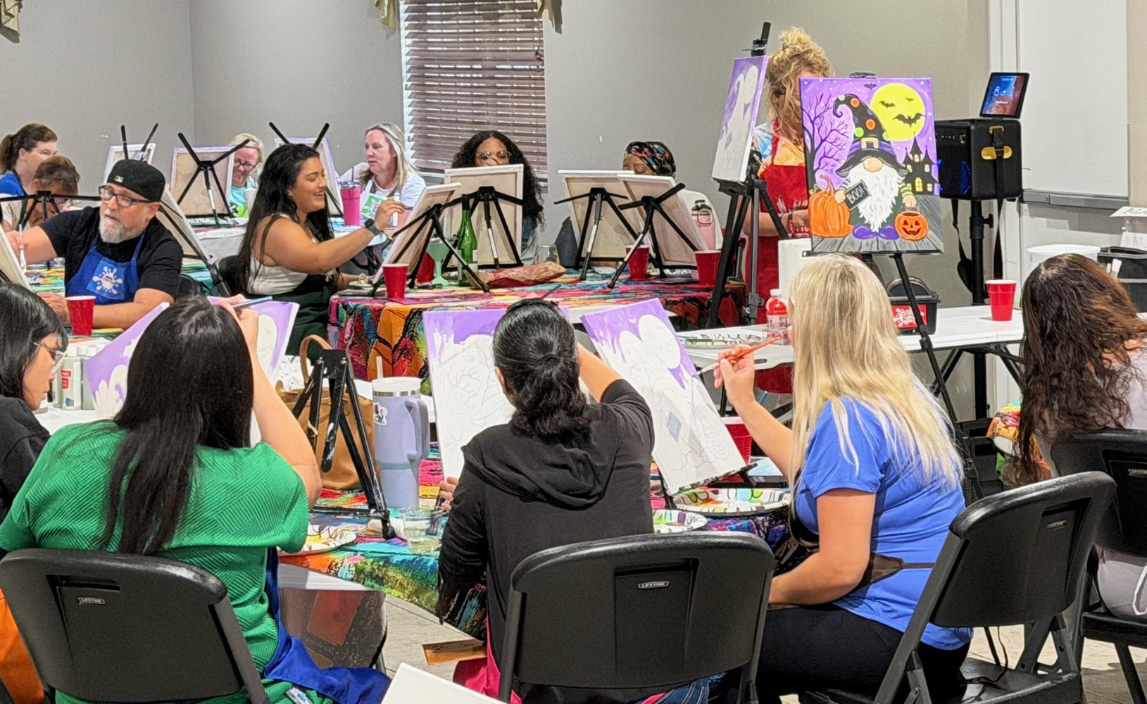 A group of people in an art class painting Halloween-themed pictures. Many are seated at tables with canvases, and one person is standing, working on their canvas. The room is decorated with colorful artwork and art supplies, with some paintings of w