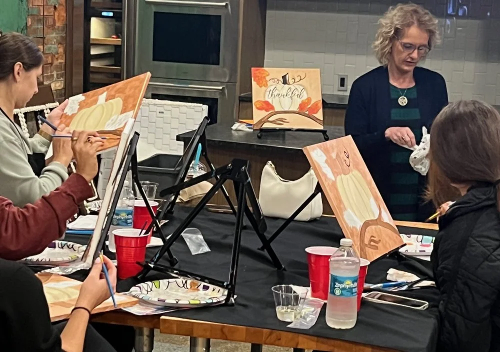 A group of people participating in a painting class, working on fall-themed artwork featuring pumpkins and autumn leaves. An instructor guides the class, and painting supplies including canvases, brushes, and water bottles are visible on the table.